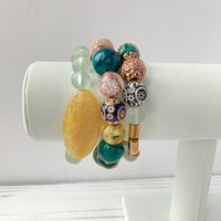 A set of three bracelet stack with green, blue, yellow, and gold beads. The bracelets are displayed on a white stand.