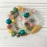 Stretch bracelet set of three bracelets with green, pink, gold, and blue beads. The bracelets are made of glass and metal beads and are arranged in a circle on a white wooden surface. Stacking bracelets for women. Lenora Dame bracelets.