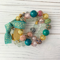 A multi-strand bracelet set with turquoise, pink, gold, and white beads. The bracelet is made of various materials including glass, metal, and wood.