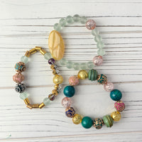 Three bracelets with different colored beads and charms. One bracelet is made of light green beads, one is made of gold and brown beads, and one is made of teal, pink, and gold beads. Stacking bracelets by Lenora Dame.