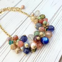 Statement necklace featuring a cascade of colorful beads in various shapes and textures. The necklace includes gold, pink, peach, green, blue, and purple beads, some with intricate patterns. The beads are clustered together, creating a bohemian style. The necklace has a gold chain clasp.