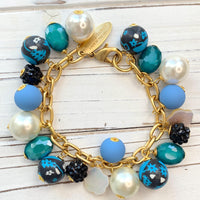 Lenora Dame jewelry. Blue and white beaded bracelet with assorted beads wire wrapped on a gold chain with lobster clasp. Black floral fimo beads, oval faceted glass beads, faux white pearls, blue rubberized beads, bumpy black beads and mother of pearl flower beads.