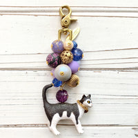 Cute bag charm for handbag with a mix of purple and gold beds on a gold chain and a black and white cat with chain collar hanging at the bottom. Lenora Dame bag charm. Beaded purse charm.