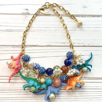 A whimsical statement necklace features a gold chain adorned with colorful beads, pearls, and miniature dinosaur figurines. The beads include shades of pink, orange, teal, blue, and purple, creating a vibrant and playful aesthetic. The necklace showcases a fun, eclectic mix of textures and materials, making it a unique accessory.