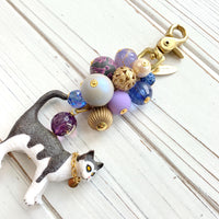 Cute bag charm for handbag with a mix of purple and gold beds on a gold chain and a black and white cat with chain collar hanging at the bottom. Lenora Dame bag charm. Beaded purse charm.