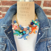 A whimsical statement necklace features a gold chain adorned with colorful beads, pearls, and miniature dinosaur figurines. The beads include shades of pink, orange, teal, blue, and purple, creating a vibrant and playful aesthetic. The necklace showcases a fun, eclectic mix of textures and materials, making it a unique accessory.