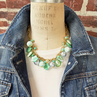 A mix of light green and light blue beads, pearls, and mother of pearl flowers hand from a gold chain necklace shown on dress form wearing a white shirt and dark blue denim jacket with a red brick wall background. Green beaded necklace for women made in the USA. Lenora Dame necklace.