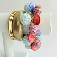 Two gold bangle bracelets and a chunky charm bracelet are shown stacked together on a white bracelet display arm. Chunky charm bracelet with assorted mix of light blue seed bead balls, floral fimo beads, blue and orange mother of pearl disc bead hung on a gold bracelet chain with lobster clasp and hangtag that says Lenora Dame Made in the USA. Lenora Dame jewelry made in USA.