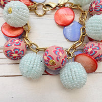 Chunky charm bracelet with assorted mix of light blue seed bead balls, floral fimo beads, blue and orange mother of pearl disc bead hung on a gold bracelet chain with lobster clasp and hangtag that says Lenora Dame Made in the USA. Lenora Dame jewelry made in USA.
