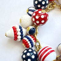 Patriotic charm bracelet with a mix of red, white and blue beads. Gold bracelet chain with lobster clasp and a Lenora Dame hang tag features blue and red round polka dot beads, and red, white and blue striped beads. Lenora Dame bracelet made in the USA.