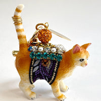 Cute cat bag charm with an orange tabby cat wearing purple and black ribbon trim embellished with green rhinestones, crystal rhinestones, and pearls. An orange rhinestone bead sits on the cat's back and is attached to the gold trigger clasp with Lenora Dame Made in USA hangtag. Lenora Dame bag charm.