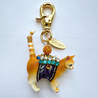 Cute cat bag charm with an orange tabby cat wearing purple and black ribbon trim embellished with green rhinestones, crystal rhinestones, and pearls. An orange rhinestone bead sits on the cat's back and is attached to the gold trigger clasp with Lenora Dame Made in USA hangtag. Lenora Dame bag charm.