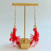 Red lobster dangle earrings for women with soft resin lobsters hanging by the tail from a chain made with 4 linked pearls, hung on French ear wires. Earrings are shown hung on a gold T-shaped earring holder. Cute lobster earrings. Lenora Dame jewelry.