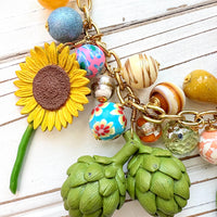 Colorful vegetable necklace for women with colorful beads and vegetable charms that include corn, artichokes, broccoli, carrots, and a sunflower hung on a gold chain along with a mix of colorful round beads. Gold chain included a hang tag that says Lenora Dame, Handmade in USA. Wacky necklace for women with vegetable charms shown on a white washed board. Lenora Dame necklace made in the USA.