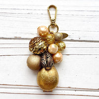 Gold tone beaded bag charm with a etched brass egg shaped hinged photo locket below a mix of pearls, lampworked glass beads, a gold sequin beads, and a black bead with gold scroll design. The beads hang on a gold chain with a gold clasp and Lenora Dame Made in USA hang tag.