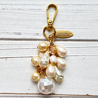 Pearl bag charm shown on a whitewashed board. A mix of cream and white faux pearls in different shapes are wire wrapped to a gold chain with gold clasp and oval hang tag that says Lenora Dame Made in USA. Lenora Dame bag charm.