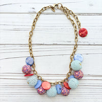 Chunky charm necklace with assorted mix of light blue seed bead balls, floral fimo beads, blue and orange mother of pearl disc bead hung on a gold necklace chain with lobster clasp and hangtag that says Lenora Dame Made in the USA. Lenora Dame necklace.