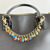 Glass Millefiori floral beads dangling from a gold chain with a trigger clasp on either end attached to the straps of a black leather handbag. Floral glass beads purse accessory. Lenora Dame bag charm.