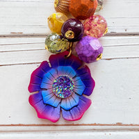 Colorful beaded bag charm filled with assorted beads in different shapes in vibrant colors. A large pink, purple and blue flower is at the bottom of the bead cluster. A gold trigger clasp is above the beads. Lenora Dame bag charms.