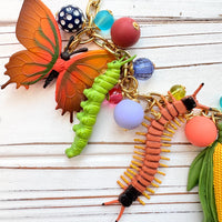 Garden bug necklace for women, with a mix of round beads, ant, butterfly, caterpillar, centipede, ladybug, corn and carrot figurines hung on a gold chain shown on a white board background. Bug jewelry for women. Lenora Dame necklace made in the USA.