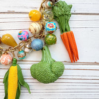 Colorful vegetable necklace for women with colorful beads and vegetable charms that include corn, artichokes, broccoli, carrots, and a sunflower hung on a gold chain along with a mix of colorful round beads. Gold chain included a hang tag that says Lenora Dame, Handmade in USA. Wacky necklace for women with vegetable charms shown on a white washed board. Lenora Dame necklace made in the USA.