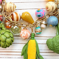 Colorful vegetable necklace for women with colorful beads and vegetable charms that include corn, artichokes, broccoli, carrots, and a sunflower hung on a gold chain along with a mix of colorful round beads. Gold chain included a hang tag that says Lenora Dame, Handmade in USA. Wacky necklace for women with vegetable charms shown on a white washed board. Lenora Dame necklace made in the USA.