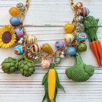 Colorful vegetable necklace for women with colorful beads and vegetable charms that include corn, artichokes, broccoli, carrots, and a sunflower hung on a gold chain along with a mix of colorful round beads. Gold chain included a hang tag that says Lenora Dame, Handmade in USA. Wacky necklace for women with vegetable charms shown on a white washed board. Lenora Dame necklace made in the USA.