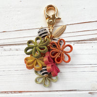 Floral bag charm with four mesh metal flowers in fall colors of green, orange and yellow, black and white striped beads with rhinestones and pumpkin orange pearls. Gold trigger clasp at the top with a gold oval charm that says Lenora Dame Made in USA.