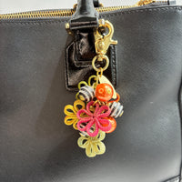 Floral bag charm with four mesh metal flowers in fall colors of green, orange and yellow, black and white striped beads with rhinestones and pumpkin orange pearls. Gold trigger clasp at the top with a gold oval charm that says Lenora Dame Made in USA. Lenora Dame bag charms shown on the strap of a black leather handbag.