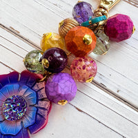 Colorful beaded bag charm filled with assorted beads in different shapes in vibrant colors. A large pink, purple and blue flower is at the bottom of the bead cluster. A gold trigger clasp is above the beads. Lenora Dame bag charms.