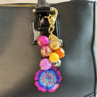 Colorful beaded bag charm filled with assorted beads in different shapes in vibrant colors. A large pink, purple and blue flower is at the bottom of the bead cluster. A gold trigger clasp is above the beads. The bag charm is shown hung on a black leather handbag strap. Lenora Dame bag charm.