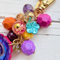 Colorful beaded bag charm filled with assorted beads in different shapes in vibrant colors. A large pink, purple and blue flower is at the bottom of the bead cluster. A gold trigger clasp is above the beads. Lenora Dame bag charms.
