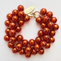 A gold chain bracelet with multiple strands of shiny rust orange faux pearls. The bracelet has a small tag that reads 'Lenora Dame' and 'Made in the USA.'