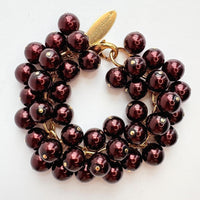 A gold chain bracelet with multiple strands of shiny dark burgundy faux pearls. The bracelet has a small tag that reads 'Lenora Dame' and 'Made in the USA.'