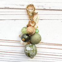 Beaded bag charm with a mix of bead styles in shades of green, gold, and pearl. One larger round bead in a army green and white toile print hangs at the bottom and a gold trigger clasp on top of all of the beads. Brass oval hang tag near the trigger clasp that says "Lenora Dame Handmade in USA". Lenora Dame bag charm.