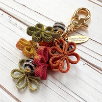 Floral bag charm with four mesh metal flowers in fall colors of green, orange and yellow, black and white striped beads with rhinestones and pumpkin orange pearls. Gold trigger clasp at the top with a gold oval charm that says Lenora Dame Made in USA.