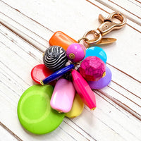 Colorful beaded bag charm with a mix of different shaped beads in a cluster under a gold trigger clasp with a gold oval charm that says Lenora Dame Made in USA. One round black and white zebra bead is among the beads. Lenora Dame bag charm.