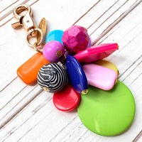 Colorful beaded bag charm with a mix of different shaped beads in a cluster under a gold trigger clasp with a gold oval charm that says Lenora Dame Made in USA. One round black and white zebra bead is among the beads. Lenora Dame bag charm.