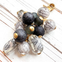 Black and white chunky bag charm with irregular shaped Lucite clear beads with a newspaper print inside hang with round black foil beads and black glass beads in a cluster under a gold trigger clasp. Lenora Dame bag charm.