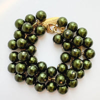 A gold chain bracelet with multiple strands of shiny hunter green faux pearls. The bracelet has a small tag that reads 'Lenora Dame' and 'Made in the USA.'