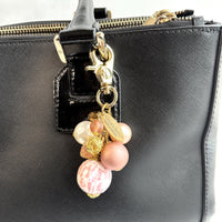 Beaded bag charm with a mix of bead styles in shades of pink, peach, gold and pearl. One larger round bead in a pink and white toile print hangs at the bottom and a gold trigger clasp on top of all of the beads. Brass oval hang tag near the trigger clasp that says "Lenora Dame Handmade in USA". Lenora Dame bag charm.