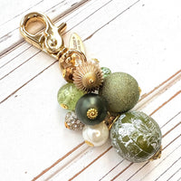 Beaded bag charm with a mix of bead styles in shades of green, gold, and pearl. One larger round bead in a army green and white toile print hangs at the bottom and a gold trigger clasp on top of all of the beads. Brass oval hang tag near the trigger clasp that says "Lenora Dame Handmade in USA". Lenora Dame bag charm.