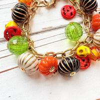 A gold chain bracelet with a variety of charms including green, orange, and white pumpkin charms, a skull charm, and a tag that reads Lenora Dame Made in the USA. Cute pumpkin charm bracelet.