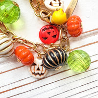 A gold chain bracelet with a variety of charms including green, orange, and white pumpkin charms, a skull charm, and a tag that reads Lenora Dame Made in the USA. Cute pumpkin charm bracelet.