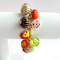 A gold chain bracelet with a variety of charms including green, orange, and white pumpkin charms, a skull charm, and a tag that reads Lenora Dame Made in the USA. Cute pumpkin charm bracelet.