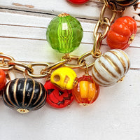 A gold chain bracelet with a variety of charms including green, orange, and white pumpkin charms, a skull charm, and a tag that reads Lenora Dame Made in the USA. Cute pumpkin charm bracelet.
