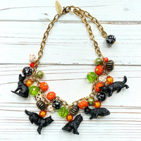 A gold chain necklace with black cat charms, orange, green, and white pumpkin charms, and black and white beads. The necklace is on a white wooden background. Lenora Dame cat necklace. Cute necklace for Halloween.