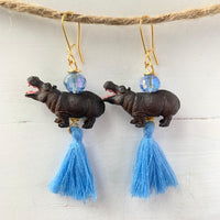 Hippo earrings with miniature brown hippopotamus, topped with a small faceted light blue bead, and blue thread tassels hanging below. Earrings have gold ear wires.
