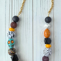Lenora Dame Halloween necklace with a mix of oversized beads strung together and attached to gold-plated cable chain with lobster clasp. The large round beads include black foil texture, purple seed bead balls, yellow spacers, black squares and wooden balls decoupaged n a mix of vintage textile prints. A turquoise colored carved sugar skull with orange and gold star eyes is strung among the beads on the left side.