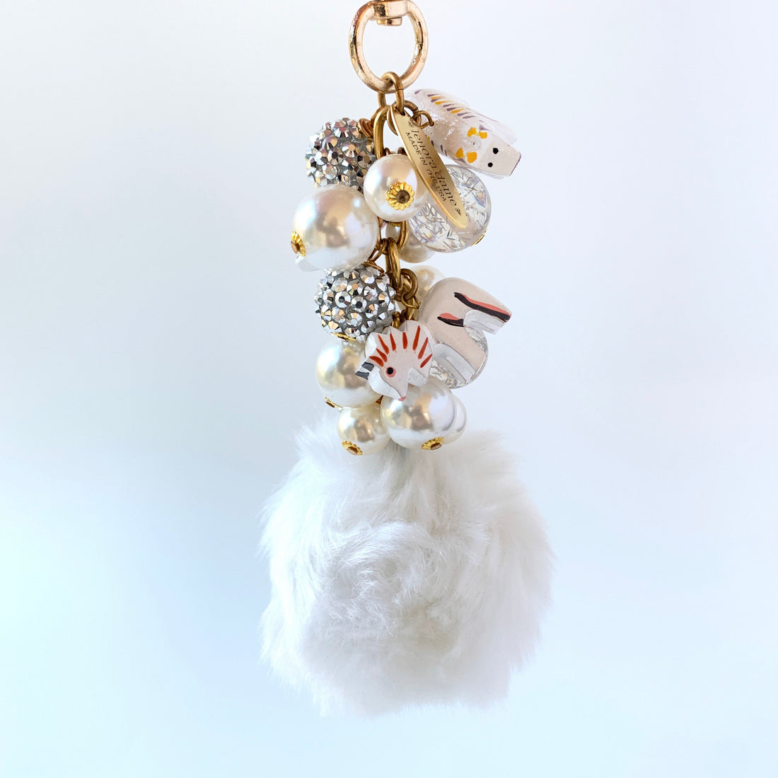 A white fluffy pom pom keychain with a gold chain and a variety of white, gold, and silver beads and charms. Lenora Dame bag charms.